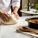 Chef’s hands kneading dough on a floured marble counter with a bubbling braiser and glossy sauce, herbs and wine in the softly blurred background.