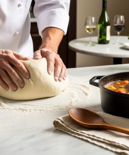 Chef’s hands kneading dough on a floured marble counter with a bubbling braiser and glossy sauce, herbs and wine in the softly blurred background.